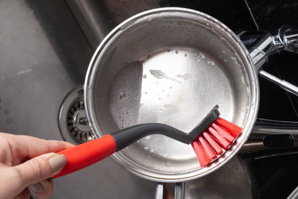 Foto de uma panela redonda de aço inox sendo lavada com uma escova redonda preta e vermelha.