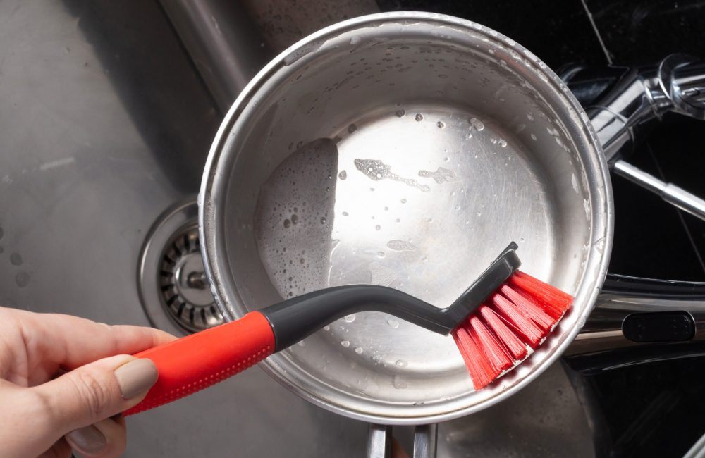 Foto de uma panela redonda de aço inox sendo lavada com uma escova redonda preta e vermelha.